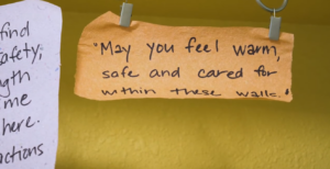 Photo of orange paper hanging on yellow wall saying "May you feel warm, safe and cared for within these walls."